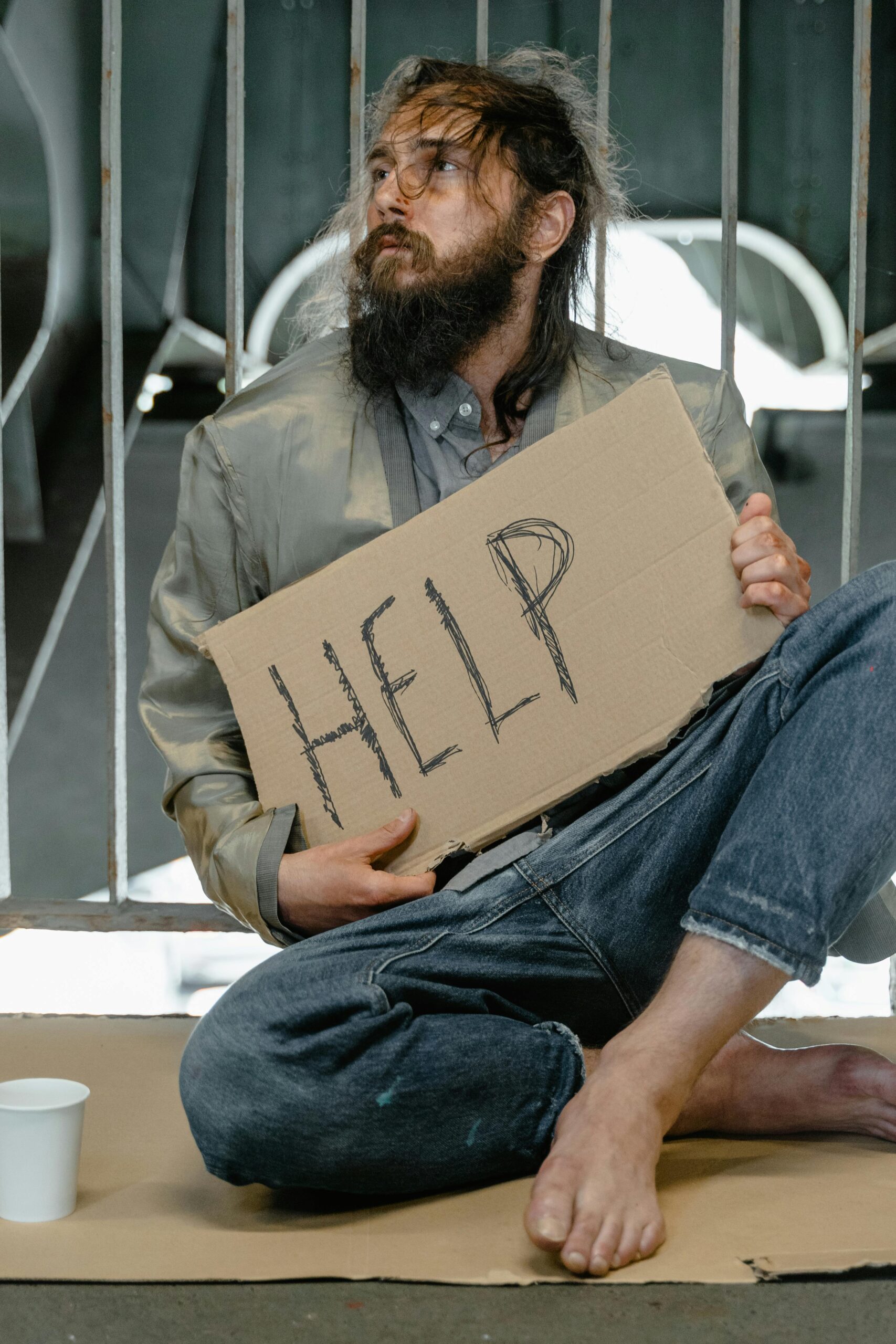 An adult man sits outdoors holding a cardboard sign with 'HELP', signifying homelessness and need.